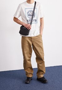 White graphic t-shirt with blue design, casual khaki pants, and black sneakers. Black crossbody bag with a zipper and logo detail.