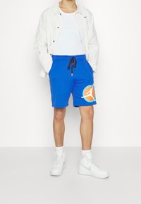 Pantaloni sportivi blu con un logo grafico arancione e bianco, dotati di una vita con cordino e di un tessuto testurizzato. Indossati con una canottiera bianca e una giacca.