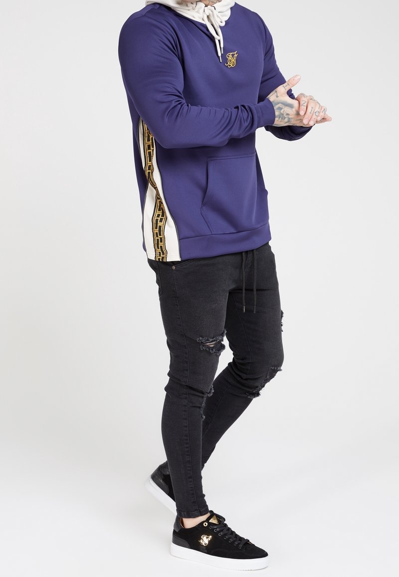 Sweat à capuche violet avec accents blancs et logo doré, associé à un jean skinny noir déchiré et des baskets noires à semelles blanches.