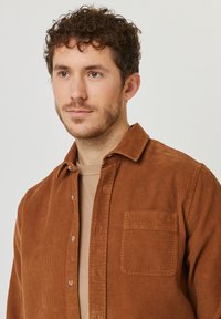 Chemise en velours côtelé marron avec un design à boutonnière, dotée d'une poche sur la poitrine gauche et d'une surface texturée en côtes. Portée par-dessus un haut beige.