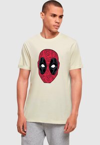 Beige bomulds T-shirt med et rødt og sort comic-stil maske grafik foran, omkranset af et blomstermønster.