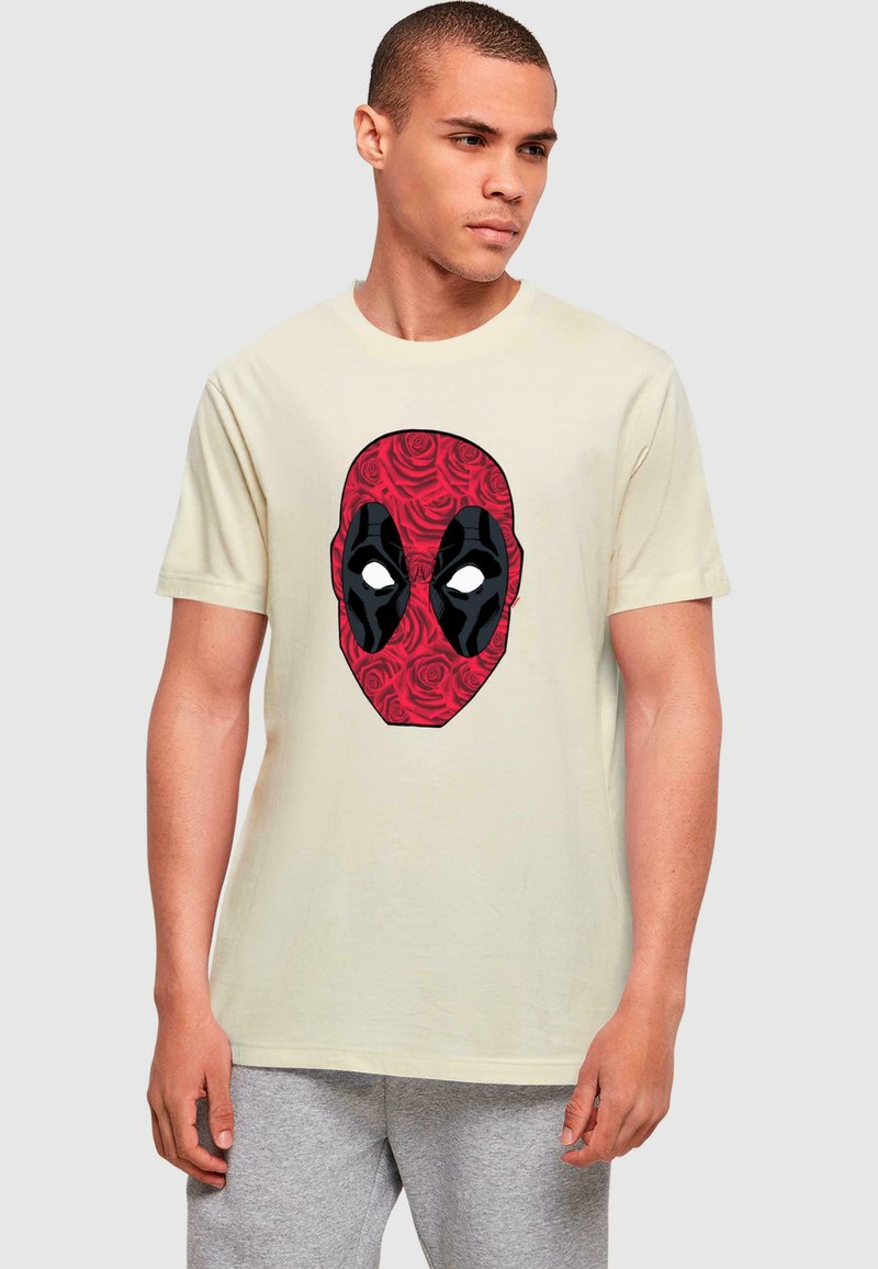 Beige bomulds T-shirt med et rødt og sort comic-stil maske grafik foran, omkranset af et blomstermønster.