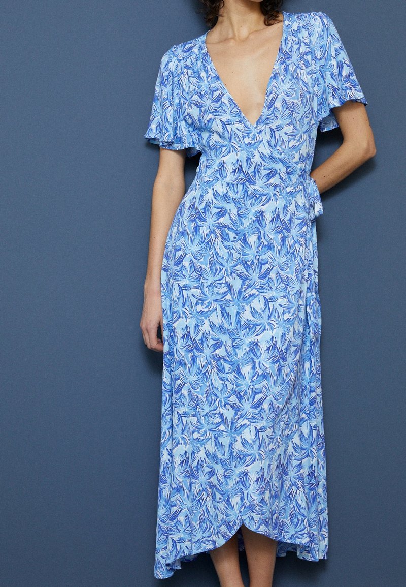 Femme portant une robe portefeuille fleurie bleu et blanc avec de courtes manches volantées sur un fond bleu uni.
