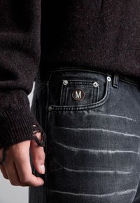 Sötétszürke farmernadrág, amely kopott hatású, finom fehér csíkokkal és egy "M" emblémával ellátott bronz gombbal rendelkezik. Egy kötött bordó pulóver látható.