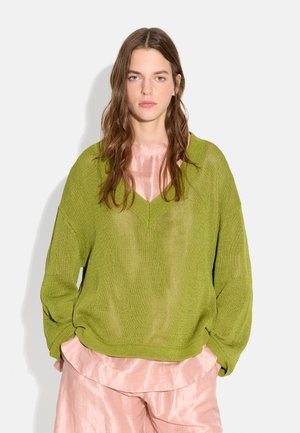 Młoda kobieta o długich brązowych włosach nosi luźny zielony sweter z dzianiny na jasnoróżowej satynowej bluzce i dopasowanych spodniach, stoi na białym tle.