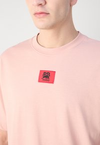 T-shirt rose clair à manches courtes en coton, ornée d'un patch rouge brodé en forme de carré avec du texte et un design noir sur la poitrine.
