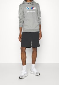 Sudadera con capucha gris con logo de BMW, cordón a juego y bolsillo tipo canguro. Pantalones cortos negros y zapatillas blancas con detalles plateados completan el conjunto.