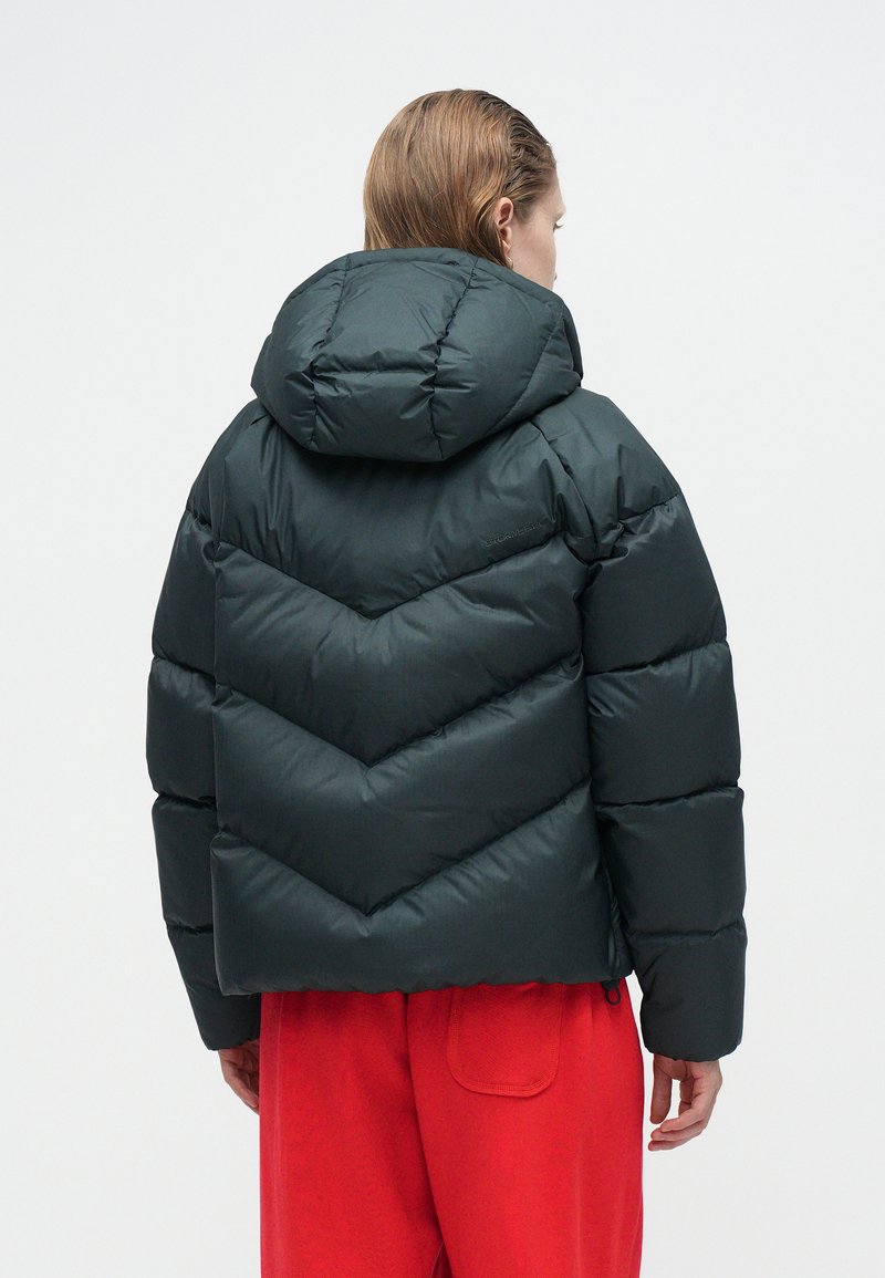 Dark green puffer jacket featuring a hood, chevron design, quilted texture, and elastic cuffs. Worn with loose red pants.