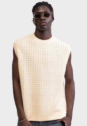 Mladý muž v bezrukávovom béžovom pletenom svetri s malým vzorom dierok, tmavých slnečných okuliaroch, náramku a sivých nohaviciach na jednoduchom pozadí.