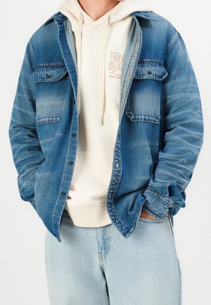 Person wearing light blue denim jacket over cream hoodie and light blue jeans, hands in pockets, against plain white background.