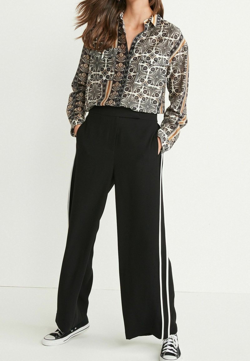Femme portant une chemise à manches longues à motifs boutonnée, un pantalon noir à jambes larges avec des bandes blanches sur les côtés, et des baskets noires avec des lacets blancs.