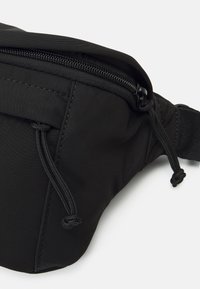 Svart tygfannypack med dragkedja, mjukt material, justerbar rem och dragsko-dekor på sidorna.