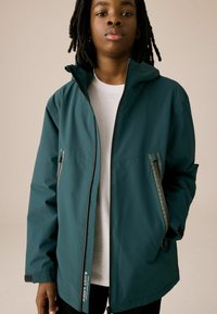 Veste imperméable teal avec fermeture éclair, capuche et deux poches latérales zippées ; présente des détails jaunes contrastants sur les fermetures éclair.