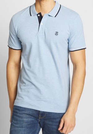 Homme portant un polo bleu clair à manches courtes avec un liseré bleu marine et un jean bleu foncé, debout avec la main droite détendue le long du corps.