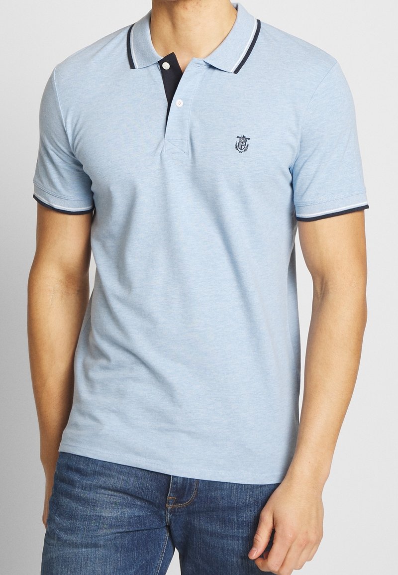 Homme portant un polo bleu clair à manches courtes avec un liseré bleu marine et un jean bleu foncé, debout avec la main droite détendue le long du corps.