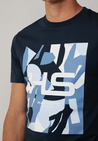 Marineblauwe T-shirt met korte mouwen, voorzien van een geometrisch lichtblauw en wit grafisch ontwerp en het logo "HACKETT SPORT" in het midden.