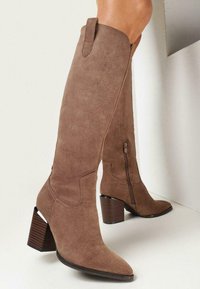 Bottes en suede marron arrivant au genou, avec un bout pointu, un talon plat, une fermeture éclair latérale et des coutures décoratives. Tige texturée et talon de motif bois.