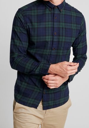 Homme portant une chemise à carreaux à boutons vert foncé et bleu marine et un pantalon beige, ajustant la manche avec ses mains.