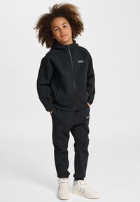 Svart zip-up hoodie och matchande byxor i mjukt tyg, med sidofickor och en subtil logo på bröstet och låret.