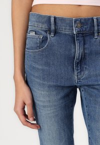 Jeans de mezclilla azul con una cintura de media altura, bolsillo delantero, herrajes plateados y un ligero desvanecimiento. Textura suave con un corte recto clásico.