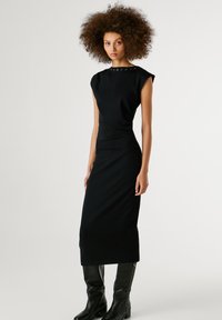 Abito midi aderente nero con maniche corte, scollatura rotonda e borchie decorative sulle spalle. Indossato con stivali neri al ginocchio.