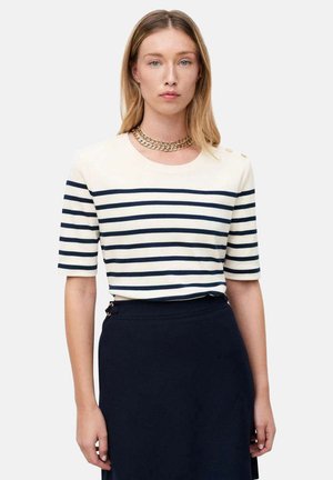 Femme aux cheveux blonds lisses portant un haut à manches courtes rayé blanc et bleu marine, un collier chaîne en or, et une jupe unie bleu marine foncé.