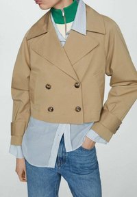 Cappotto trench beige sfumato con bottoni doppi, revers appuntiti e design strutturato su una camicia a righe blu e jeans.