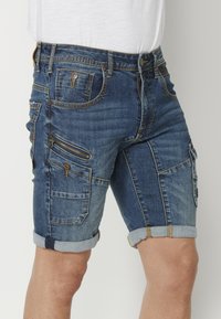 Denim kravu šorti, zilā krāsā ar nolietotu apdari, ar vairākiem kabatām, ar rāvējslēdzēju sānu kabatu un ruļļveida apakšmalām, lai radītu ikdienišķu izskatu.
