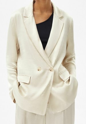 Blazer de satén beige con cierre de botones cruzados, solapas cortadas y dos amplios bolsillos frontales. Textura suave, ajuste relajado.