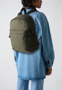 Mochila de nailon verde con forma redondeada, bolsillo frontal con cremallera y correas ajustables, llevada sobre una chaqueta de mezclilla azul y pantalones a juego.