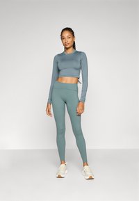 Långärmad cropped topp och högmidjade leggings i dämpad teal, tillverkade av slät, stretchig tyg. Sneakers med kräm- och bruna accenter.