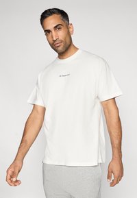 Camiseta branca de algodão com mangas curtas, decote redondo e detalhe em texto no peito. Combinada com calças de fato de treino cinzentas, criando um visual casual.