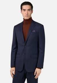 Chaqueta de traje a cuadros en color azul marino con cierre de dos botones, solapas de muesca y bolsillos frontales. Combinada con un suéter de cuello alto marrón y un pañuelo de bolsillo estampado.