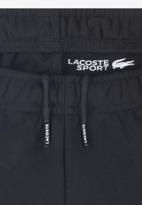 Mustad spordipüksid, millel on elastne vöökoht ja nöörid. "Lacoste Sport" bränd on siseruumis valge värviga nähtav.