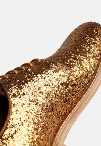 Scarpa dorata scintillante con superficie testurizzata, punta arrotondata e suola di colore chiaro. Presenta lacci marroni e una fodera interna liscia.