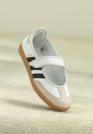 Biały i beżowy sneaker z czarnymi paskami, paskiem na rzep oraz gumową podeszwą unoszący się nad teksturowaną beżową powierzchnią.