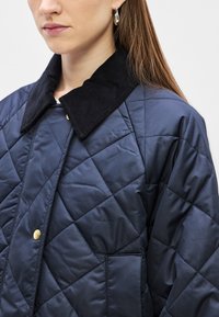 Tengerészkék steppelt dzseki fekete kordbársony gallérral, arany színű fém részletekkel és gyémánt alakú varrással. Oldalzsebes részlettel.