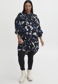 Knapparmarckad skortklänning med ett abstrakt blommönster i svart, blått och vitt. Långa ärmar och en böjd kantsömn, ihop med svarta leggings och beige stövlar.