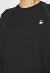 Personne portant une chemise noire texturée à manches longues avec un petit logo blanc « RAW » sur la partie supérieure gauche de la poitrine.