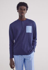 Langarmige marineblauwe shirt van een zachte stof met een blauw gestreepte pocket. Heeft een henley-halslijn met drie knoppen. Gecombineerd met blauw gestreepte broek.
