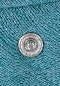 Hellblauer Strickstoff mit einem metallischen Druckknopf, der klare Gravuren am Rand zeigt und Textur sowie Detail präsentiert.