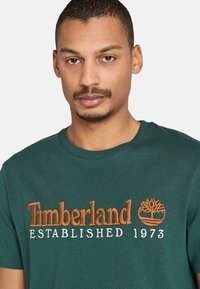 Zelené bavlněné tričko s oranžovou vyšitou grafikou loga "Timberland ESTABLISHED 1973" a stromem na hrudi. Kulatý výstřih.