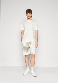 Light-colored t-shirt with a small logo, paired with patterned shorts featuring a grid design in light tones. White sneakers complete the outfit.
