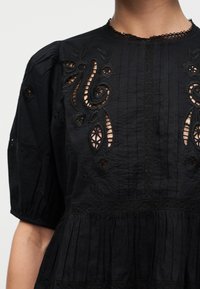 Blouse noire à manches bouffantes avec une broderie florale découpée complexe sur la poitrine et le haut des manches.