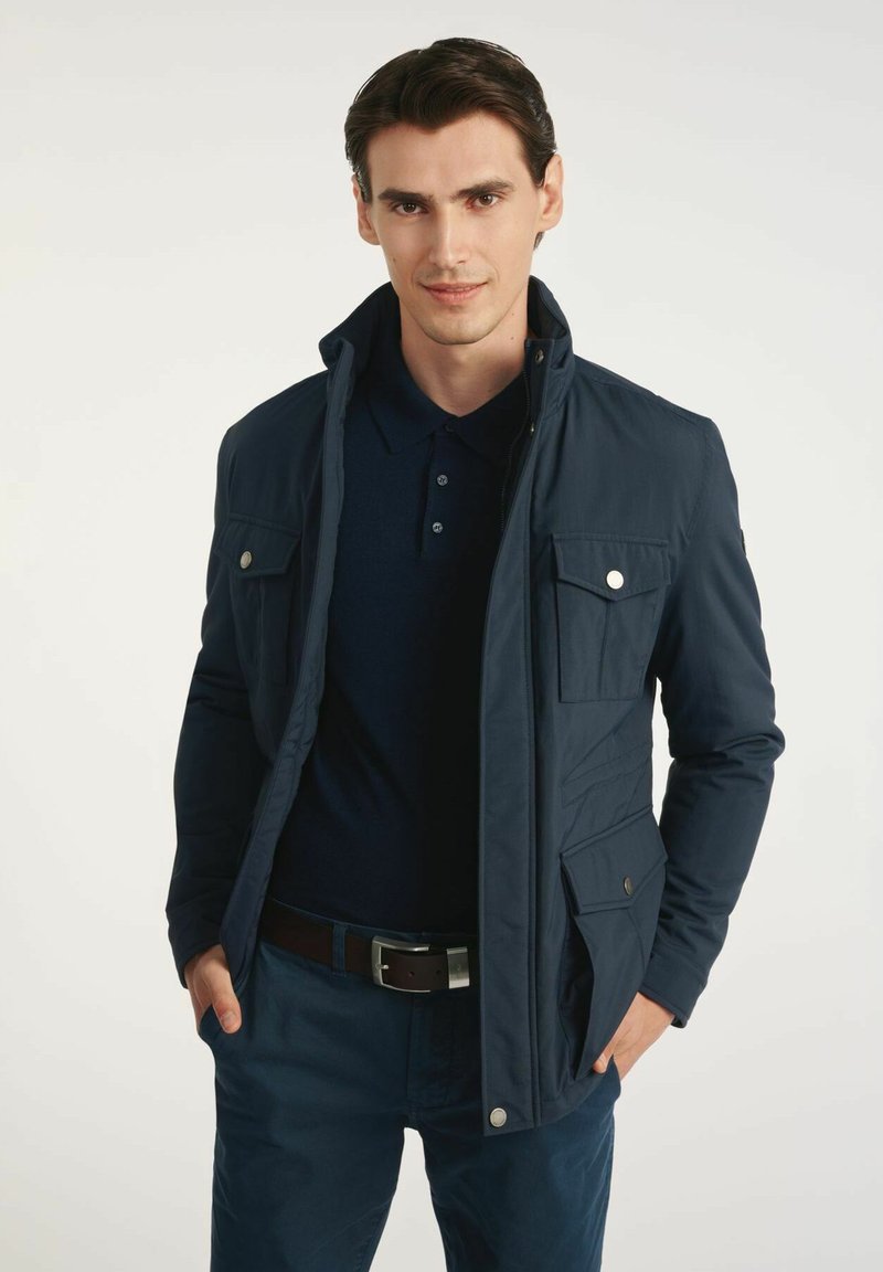 Granatowa kurtka z wodoodpornego materiału, z dwoma kieszeniami na piersi i zatrzaskami. Nosi się ją na ciemnej koszulce polo o teksturowanym wykończeniu.