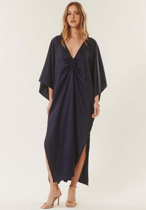 Femme portant une robe maxi ample bleu marine foncé à col en V avec un détail froncé à l'avant et des fentes latérales, associée à des sandales à talons hauts dorées.