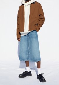 Brązowa kurtka na szarym hoodie, w połączeniu z niebieskimi dżinsowymi szortami do kolan i czarnymi sznurowanymi butami o grubych podeszwach. Widoczne białe skarpetki.