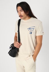 Beige bomulds T-shirt med blåt logo og tekstdesign på venstre bryst. Modellen holder en sort taske med en rem og har lyse bukser på.