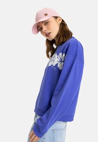 Sudadera azul con diseño floral blanco, de corte relajado y puños acanalados. Combinada con una gorra rosa clara que presenta un parche con el logotipo.