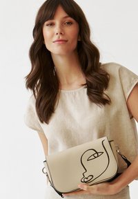 Borsa a tracolla beige con un disegno a linea nera di un volto. Materiale in pelle liscia con chiusura a patta e tracolla rimovibile.